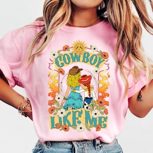 Puede incluir: Una camiseta rosa claro con un estampado gr&aacute;fico de dos figuras con cabello rubio y rojo que llevan sombreros de vaquero. El dise&ntilde;o incluye elementos florales, un sol y el texto "COWBOY LIKE ME" en letras grandes de color verde azulado.
