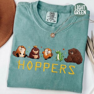 May include: Light green t-shirt with embroidered cartoon animals and the word "HOPPERS" in yellow. The shirt features a beaver, a bear, a frog, and other animals. The shirt is a size small.