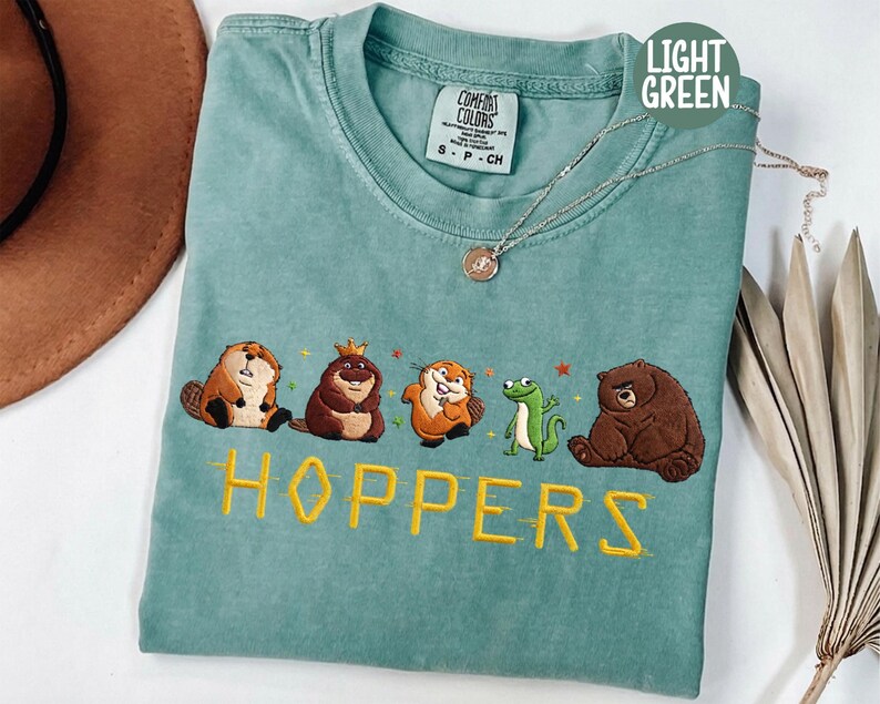 May include: Light green t-shirt with embroidered cartoon animals and the word "HOPPERS" in yellow. The shirt features a beaver, a bear, a frog, and other animals. The shirt is a size small.