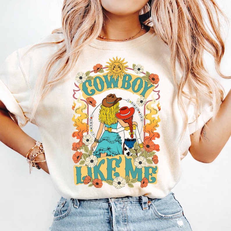 Puede incluir: Una camiseta de color crema con un estampado gr&aacute;fico de dos figuras de pelo largo, una con un sombrero de vaquero marr&oacute;n y la otra con un sombrero rojo, rodeadas de flores, un sol y llamas, con el texto COWBOY LIKE ME impreso en azul.