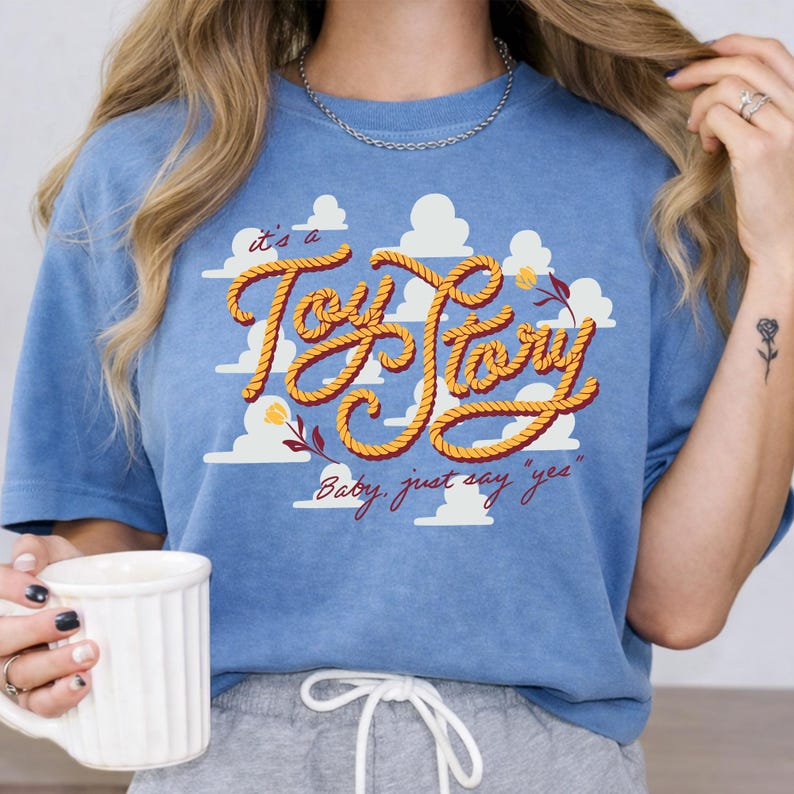 May include: A blue t-shirt featuring a graphic of white clouds and the text "it's a Toy Story" written in a yellow rope-style font. Below the main text, the phrase "Baby, just say 'yes'" is printed in red cursive script with small floral accents.