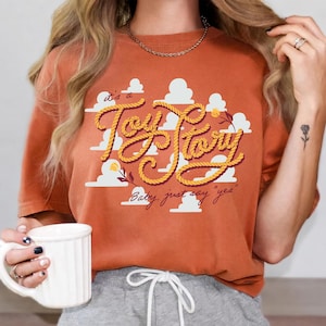 May include: A burnt orange t-shirt featuring a graphic of white clouds and the text "it's a Toy Story" written in a yellow rope-style font, with the additional script text "Baby, just say 'yes'" printed below in a dark brown cursive font.