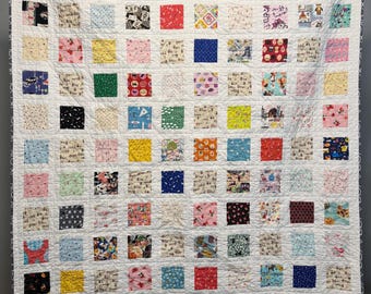 May include: A handmade quilt with a white background and numerous colorful square patches. The patches feature various patterns, including floral, geometric, and novelty prints. The quilt is displayed on a wall, held by black clips.