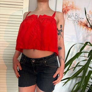 Könnte beinhalten: Ein leuchtend rotes Crop-Top mit schmalen Trägern und einer Knopfleiste vorne. Dazu werden dunkelblaue Jeansshorts getragen. Die Shorts haben einen Knopfverschluss und Taschen. Das Outfit ist lässig und stilvoll.