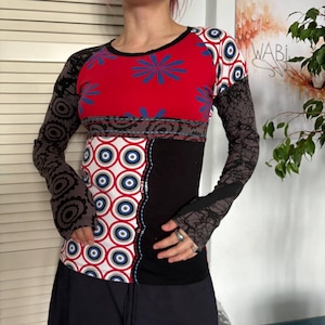 May include: A long-sleeve shirt featuring a red upper section with blue floral patterns, a white lower section with red and blue circular motifs, and a solid black panel. The sleeves are dark grey with black circular and abstract patterns.