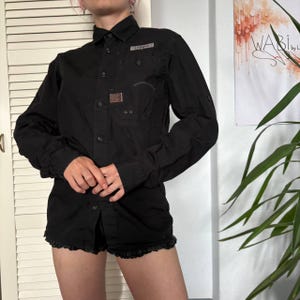 May include: A black long-sleeved button-down shirt featuring a structured collar, a chest pocket with a small rectangular patch, and a buttoned placket. The shirt has a straight hemline and long sleeves with cuffs, constructed from a solid black fabric.