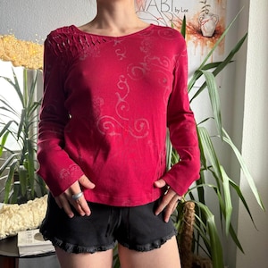 Könnte beinhalten: Ein langärmeliges rotes Oberteil mit einem dezenten, tonalen Wirbelmuster und strukturierten Raffungen an der rechten Schulter. Das Oberteil wird mit einer schwarzen Shorts kombiniert, die einen gerüschten Saum und einen sichtbaren elastischen Bund hat.