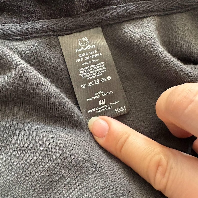 May include: Close-up of a black garment tag with the Hello Kitty logo. The tag includes size information, washing instructions, and the H&M logo. The tag is being held by a person's finger.
