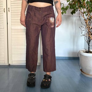 May include: Dark brown trousers featuring a mid-rise waist with a double button closure, a small metal D-ring on the front left hip, and a decorative circular graphic patch on the upper left thigh. The pants have a straight-leg cut and a subtle sheen.