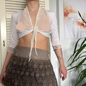May include: A white long-sleeved top with a sheer overlay tied at the front, paired with a brown skirt featuring a lace design. The outfit is a combination of textures and layers.