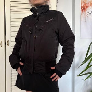 以下が含まれることがあります： ハイネックの黒いジャケットで、胸には「Bench」のブランド名がプリントされています。 ジャケットはウエストがフィットし、複数のポケットとジッパー開閉が付いています。 袖には親指用の穴があります。 ジャケットは黒いスカートと合わせて着用されています。