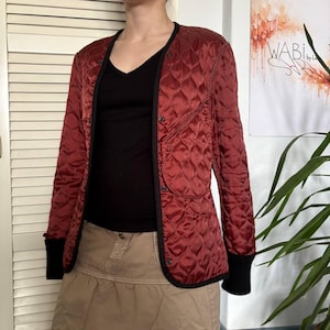 May include: A quilted jacket in a deep red hue with black trim and cuffs. The jacket features a diamond pattern and a V-neck design. It is worn over a black top and paired with a tan skirt.