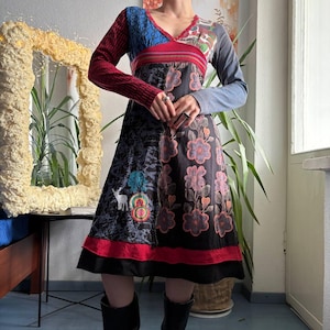 May include: A long-sleeve midi dress featuring a V-neckline and a patchwork design. The bodice combines blue, red, and grey panels. The skirt is black with a floral print, a white deer graphic, and colourful circular accents, finished with a red hem.