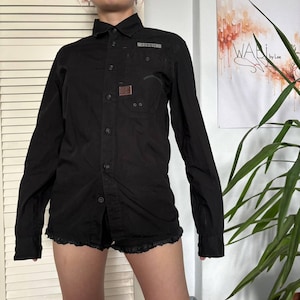 May include: A black long-sleeved button-down shirt featuring a pointed collar, a front button placket, and a single chest pocket. The shirt includes a small rectangular patch on the chest and a grey label detail above the pocket, with long sleeves.
