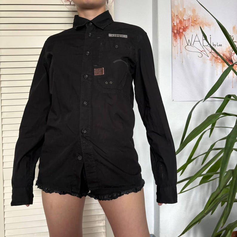May include: A black long-sleeved button-down shirt featuring a pointed collar, a front button placket, and a single chest pocket. The shirt includes a small rectangular patch on the chest and a grey label detail above the pocket, with long sleeves.
