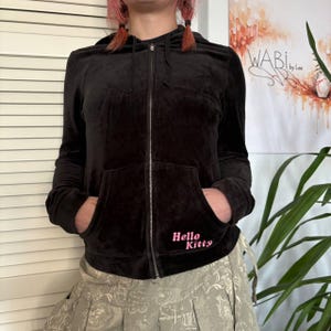 May include: A black zip-up hoodie with a front pocket and a hood. The words "Hello Kitty" are embroidered in pink on the front. The hoodie is made of a soft, velvety material.