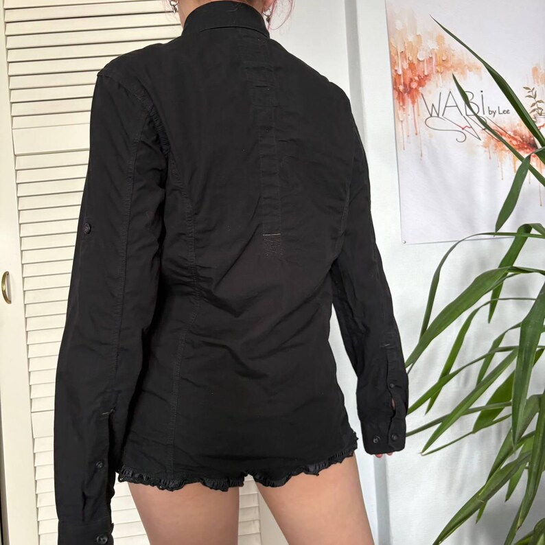 May include: A black long-sleeved button-down shirt featuring a structured collar, a vertical stitched panel down the centre back, and buttoned cuffs. The shirt has a straight hemline and is shown from the back, displaying a fitted silhouette.