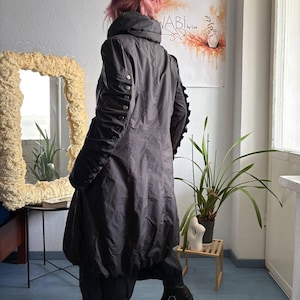May include: A dark gray, knee-length coat with a high collar and a gathered, bubble-style hem. The sleeves feature a vertical row of silver-toned metal buttons running from the shoulder to the cuff, creating a ruched effect along the arms.