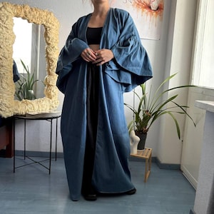 May include: A long, open-front blue duster coat featuring wide, tiered bell sleeves. The garment is worn over a black crop top and black trousers. The coat has a smooth, solid-colored fabric construction with a relaxed, flowing silhouette that extends to the floor.