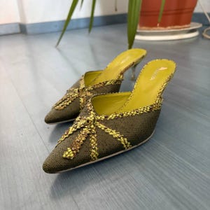 Puede incluir: Un par de mules de punta verde oliva con un patrón decorativo amarillo y marrón. Los tacones son finos y plateados. El interior es de color amarillo brillante.