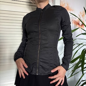 May include: A charcoal gray long-sleeve jacket featuring a high mandarin collar, a full-length front zipper closure, and vertical seam detailing that runs along the torso. The garment is fitted with long sleeves and a straight hemline.