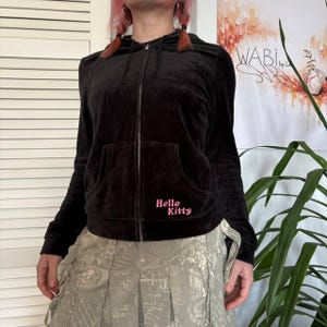 May include: Black zip-up hoodie with a front pocket and a hood. The hoodie has the words "Hello Kitty" in pink script on the lower left side. The material appears to be velvet.
