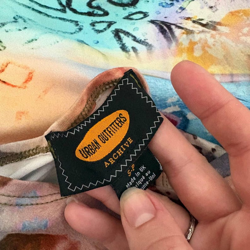 以下が含まれることがあります： Urban Outfitters Archiveの黒いタグのクローズアップ。白い文字と黄色の楕円形のロゴがあります。タグには「Urban Outfitters Archive」と「Made in UK」と書かれています。背景はカラフルな模様の生地です。