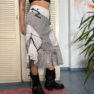 May include: A patchwork skirt with a unique design. The skirt features a mix of gray and white fabrics, including striped and patterned sections. It has a ruffled detail and decorative stitching. The skirt is paired with black boots.