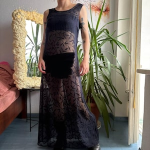 May include: A sleeveless, floor-length, sheer black dress featuring a repeating dark leaf pattern throughout the fabric. The garment has a round neckline and a solid black opaque panel across the midsection, providing coverage over the torso area.