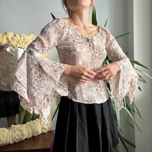 Puede incluir: Una blusa de manga larga con base blanca y estampado de cachemira marrón. La prenda tiene un escote en V con un fino detalle de cordones en el centro del pecho y unas espectaculares mangas acampanadas que se ensanchan desde el codo.