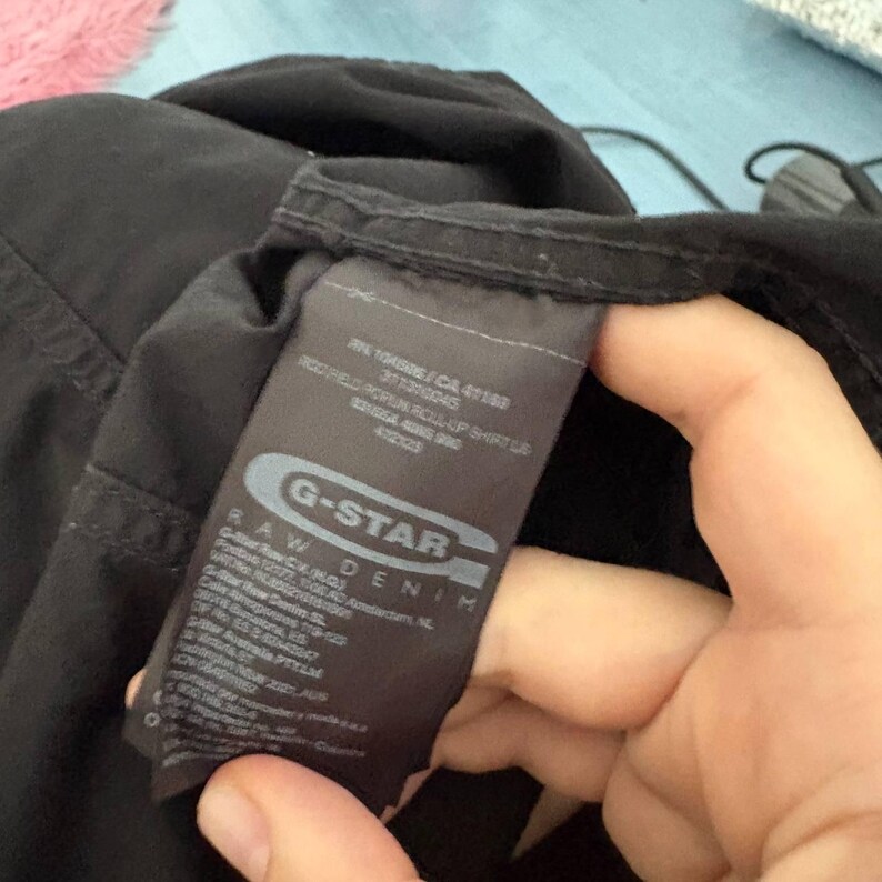 May include: A black G-Star Raw Denim shirt featuring a rectangular fabric care label. The label displays the brand logo, RN 104506, CA 41169, and the text 'ROD FIELD ROL-UP SHIRT LS'. The garment is made of black fabric with visible stitching details.