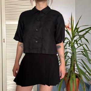 May include: A black short-sleeved button-down shirt with a classic collar and a straight hemline, paired with a black pleated mini skirt. The shirt features a front button placket with four visible dark buttons and a structured, boxy fit.