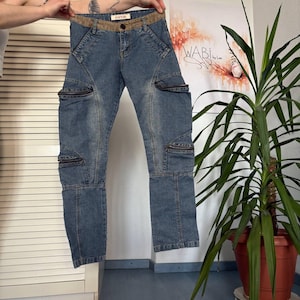 May include: Blue denim jeans with multiple zippered pockets and a brown waistband. The jeans have a unique design with angled seams and zippers on the legs. The jeans are held up by a person's hands.