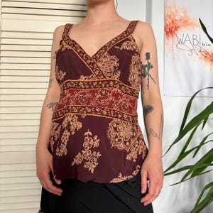 Op de afbeelding: Bordeaux tanktop met een V-hals en bloemenpatroon in beige en rood. De top heeft een decoratieve band over het midden en bandjes. De top is gemaakt van een lichtgewicht materiaal.