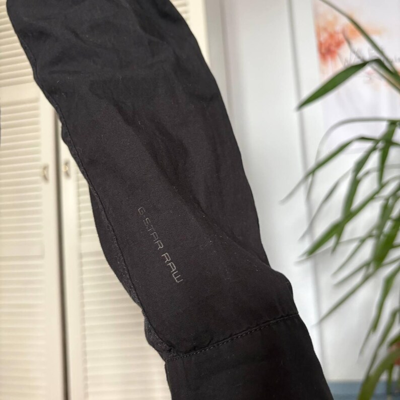 May include: A black long-sleeved garment sleeve featuring the G-STAR RAW brand name printed in a subtle, dark tone on the fabric. The sleeve includes a structured cuff at the wrist and a smooth, matte black finish across the entire visible surface.