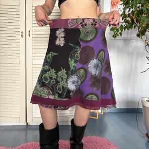 May include: A knee-length skirt featuring a split design with a black section on the left and a purple section on the right. The black side displays green bird and floral motifs, while the purple side features circular patterns. A dark maroon band borders the hem.