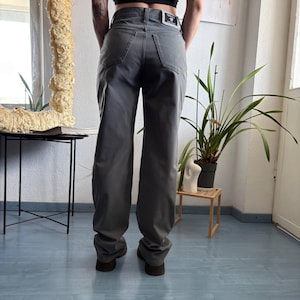 May include: Gray denim jeans featuring a straight-leg cut, two back pockets, and a rectangular Versace brand patch sewn onto the back waistband. The pants have a classic five-pocket construction with visible stitching and a button fly closure.
