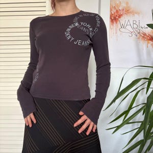 May include: A dark grey long-sleeved top featuring the text "DKNY JEANS NEW YORK CITY" in a circular design. The top has a crew neck and fitted sleeves. The model is also wearing a black and brown striped skirt.