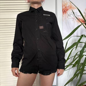 May include: A black long-sleeve button-down shirt featuring a pointed collar, a front button placket, and a chest pocket with a small rectangular patch. The shirt has a structured design with visible stitching details and a clean, solid black finish.