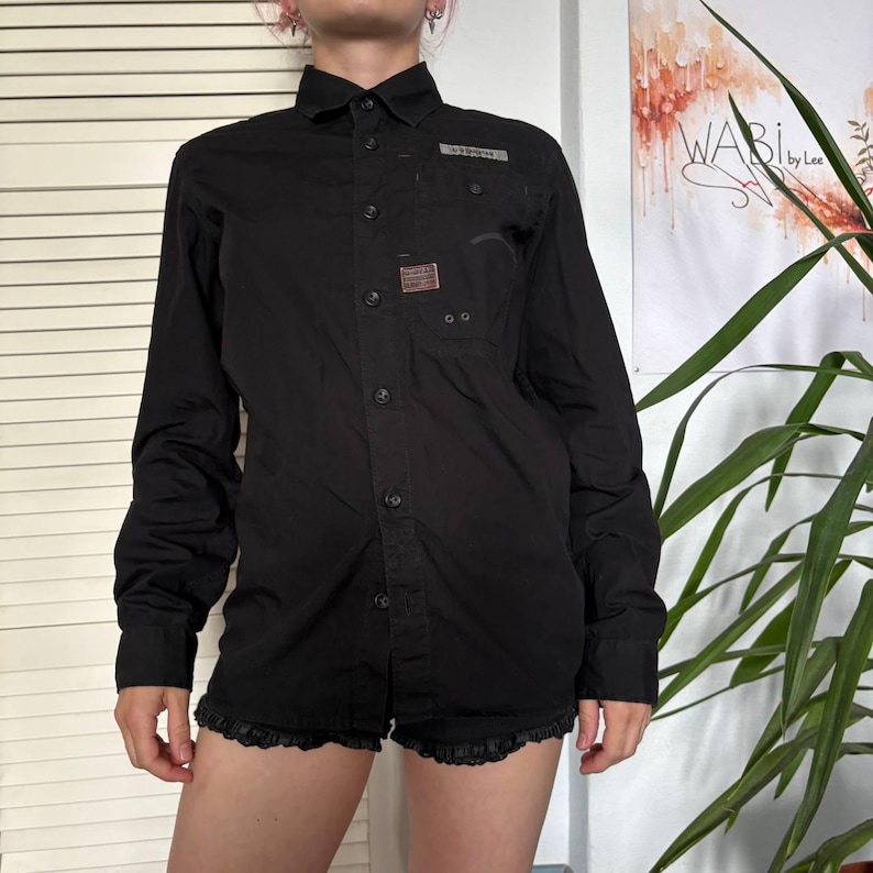 May include: A black long-sleeve button-down shirt featuring a pointed collar, a front button placket, and a chest pocket with a small rectangular patch. The shirt has a structured design with visible stitching details and a clean, solid black finish.