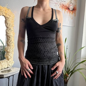 May include: A black tank top with a V-neckline and double shoulder straps. The garment features a subtle grey diamond-patterned graphic across the midsection and a textured grey band at the hem. The design includes a small white label on the side.