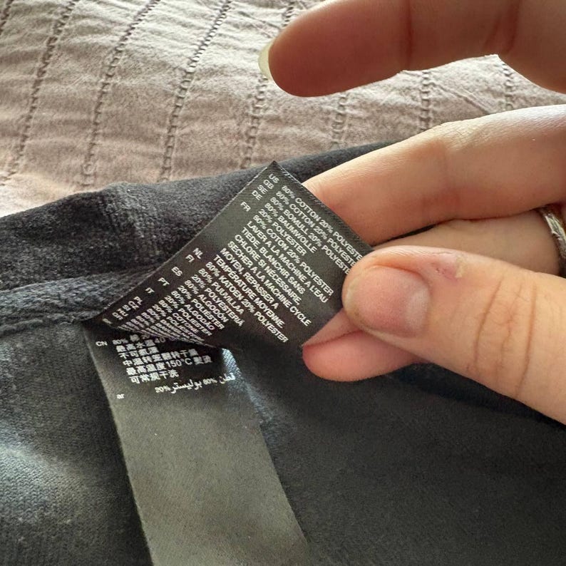 May include: Close-up of a black garment label with white text. The label lists fabric composition as 80% cotton and 20% polyester in multiple languages. Washing instructions are also included. The label is held by a hand.