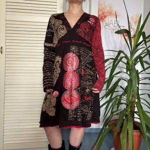 Pode incluir: Vestido preto de manga comprida com decote em V, com estampas paisley, florais e de escrita. O vestido tem detalhes vermelhos em uma manga e na bainha, com a palavra "Desigual" impressa no peito.