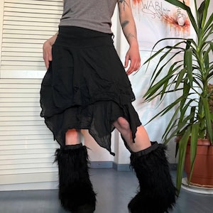 May include: Black asymmetrical skirt with a layered design. The skirt is paired with black furry leg warmers. The outfit is worn with a gray top. A plant in a brown pot is in the background.