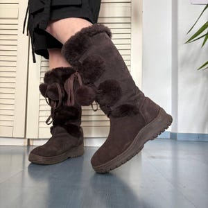 Könnte beinhalten: Braune Kniehochstiefel aus Wildleder mit Kunstfellbesatz und Bommelakzenten. Die Stiefel haben eine runde Spitze und eine dicke, strukturierte Sohle. Die Stiefel sind in einem warmen, erdigen Braunton gehalten.