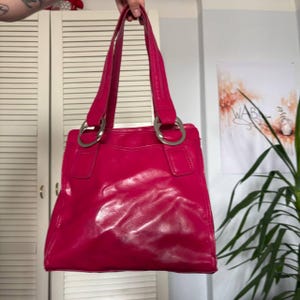 Puede incluir: Un bolso de mano color rosa fucsia brillante con un acabado lustroso similar al charol. El bolso cuenta con dos correas verticales para el hombro unidas al cuerpo principal mediante grandes anillos metálicos circulares plateados. Diseño cuadrado.