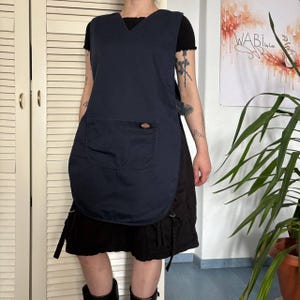 May include: A navy blue Dickies apron with a large front pocket. The apron features a V-neck and a side fastening. The Dickies logo is on the pocket. The apron is worn over a black outfit.