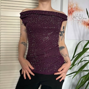May include: A dark purple off-the-shoulder top with a fitted silhouette. The top is covered in small, sparkling embellishments. The model is wearing black pants. The top is a stylish piece of apparel.