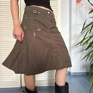 May include: A knee-length brown skirt featuring a structured waistband with a button closure, multiple silver-toned metal grommets, and reinforced stitching details. The skirt has a flared silhouette with panel construction and a smooth finish.