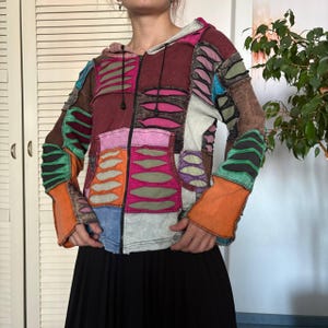 May include: A long-sleeve zip-up hoodie featuring a patchwork design with various colors including maroon, orange, blue, green, and pink. The garment includes black drawstring cords and decorative cut-out leaf-shaped patterns throughout the fabric.